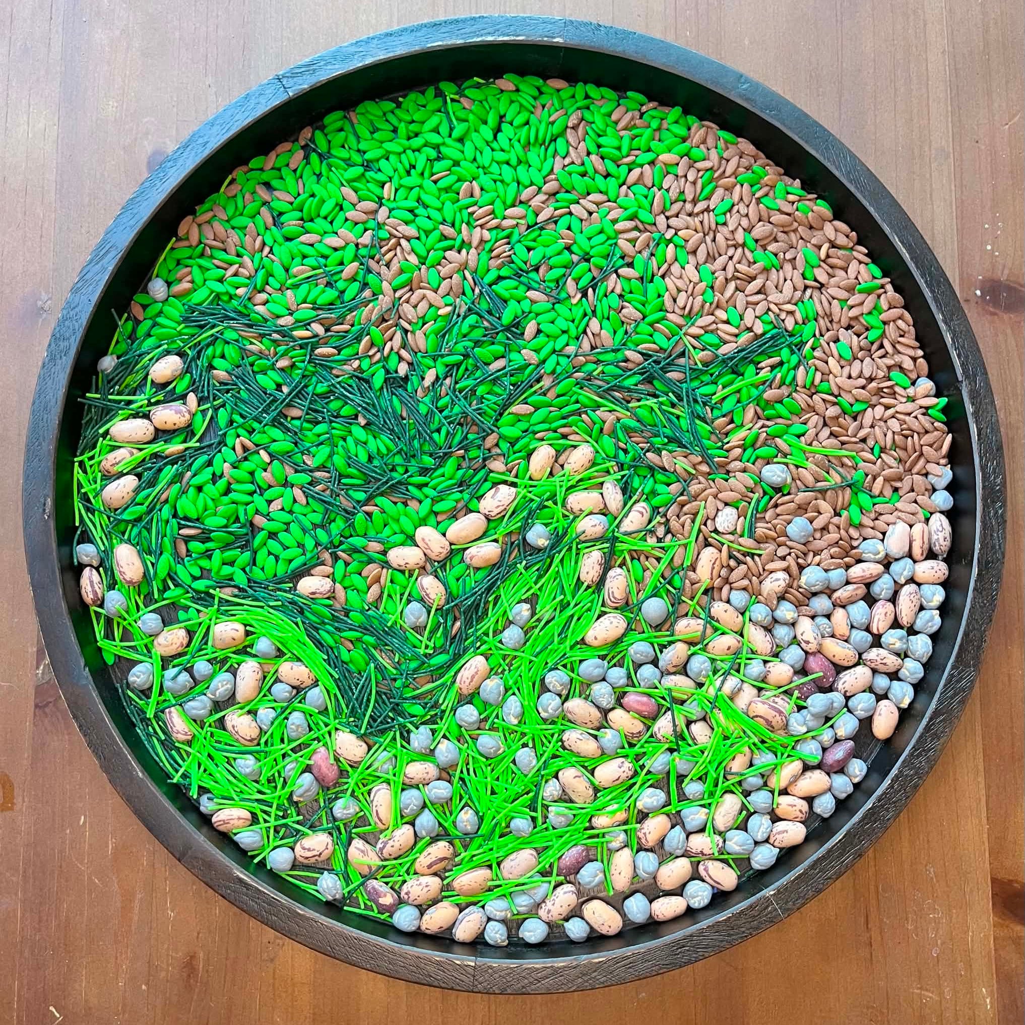 Decorative arrangement of green and multicolored seeds in a circular pattern on a wooden surface.